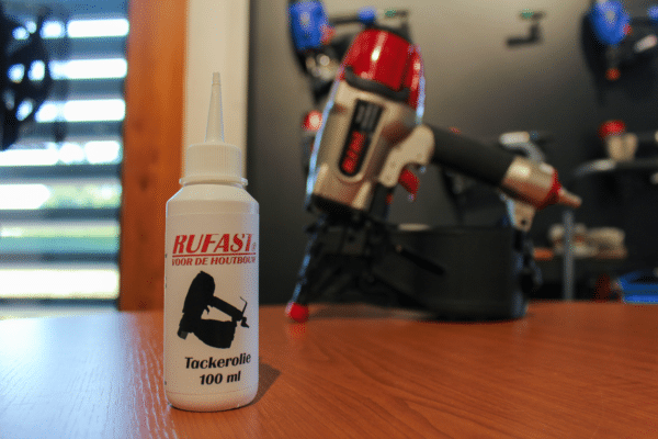 Fles Rufast-tackerolie met een pneumatische coilnailer op de achtergrond